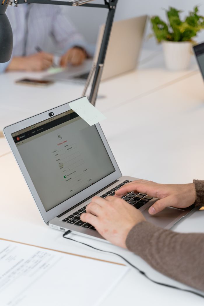 Services Close-up of hands typing on a laptop in a modern office with coworkers in the background.